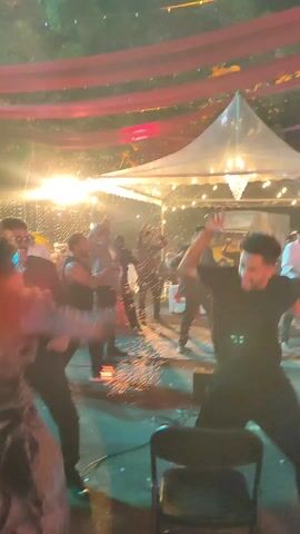 A group of people are dancing outdoors at night in a festive setting, with confetti falling. There are red drapes and string lights above, with a white tent in the background. Some of the dancers appear to be holding flaming torches. One dancer in a black outfit is particularly animated, moving a chair around. Others watch or participate in the dancing.
