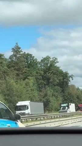 The video captures a scene on a highway, likely in Germany, given the presence of German language text on a vehicle in the foreground. The perspective is from inside a car, looking out the passenger side window. The highway appears to have multiple lanes, with traffic moving in the same direction.

In the immediate foreground, the rear passenger side of a light-colored car is visible. It has a blue and white graphic on its door with text in German that appears to be a slogan or advertisement, partially obscured. The car's side mirror is also visible.

Beyond the foreground car, the highway stretches into the distance. Several trucks are driving on the highway. One prominent truck is a red tractor unit pulling a white trailer. Further down the road, there are at least two more white tractor units pulling trailers. One trailer has "DEM" printed on its side in blue lettering.

The highway is bordered on the right by a concrete barrier system, typical of European highways. Beyond the barrier, there is a dense green forest with various types of trees, indicating a rural or semi-rural setting. The sky above is partly cloudy, with patches of blue sky visible between the clouds. The lighting suggests it is daytime, possibly late morning or afternoon, with a bright sun.

The video appears to be a continuous shot, showing the movement of traffic and the passing landscape from the viewpoint of a passenger. The overall impression is a typical highway journey through a wooded area.