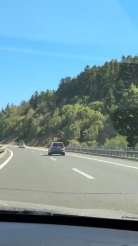 The video shows a view from inside a car driving on a highway. The road is paved with asphalt and has white lane markings. On the right side of the road, there is a steep embankment covered with green trees and vegetation. On the left side, there is a highway barrier and another lane of traffic, with trucks and cars visible in the distance. The sky is clear and blue. The car is moving forward, and the scenery is passing by.