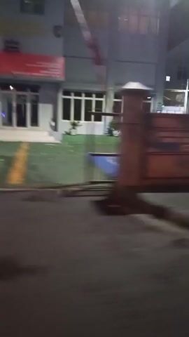 The video shows a nighttime scene from a moving vehicle, possibly a car or a bus. The camera is shaky and the footage is blurry due to motion. The view is primarily of the side of the road and buildings.

At the beginning, a dark, brick-like wall with horizontal wooden planks is visible. Above the wall, a building with several windows is dimly lit. A sign with blue and yellow lettering is affixed to the wall. The ground in front of the wall appears to be wet, with dark patches that could be puddles or stains.

As the vehicle moves, the scenery changes. A building with multiple floors and windows comes into view, illuminated by artificial lights. A street light is visible. Later, a large, brown structure with horizontal slats, resembling a garage door or a shutter, is seen. Further along, there are glimpses of other buildings with lights, including some with red and yellow elements, and a white vehicle parked. The overall impression is of driving through a dimly lit urban or suburban area at night. There are no discernible people or direct interactions visible.