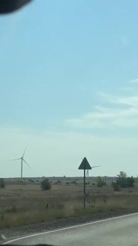 The video begins with a close-up shot of a triangular road sign on the left side of the frame. In the background, two wind turbines are visible against a pale blue sky with scattered clouds. The scene then pans to the right, revealing another road sign with two circular symbols, one above the other. The camera continues to move, showing a paved road stretching into the distance. More wind turbines are scattered across a flat, grassy landscape on the right side of the road. The sky remains a clear blue with some wispy clouds. Several cars are driving on the road, and the vegetation on the sides appears dry and sparse. The video ends with a shot of a line of wind turbines in the distance, with the road continuing towards them under a bright sky.