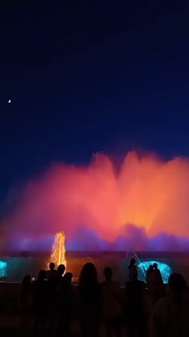 A group of people are silhouetted against a colorful fountain show at night. The fountain sprays water in arcs that are illuminated with red, orange, pink, and blue lights. The night sky is dark blue with a crescent moon visible in the upper left corner. The people appear to be standing and watching the fountain show.