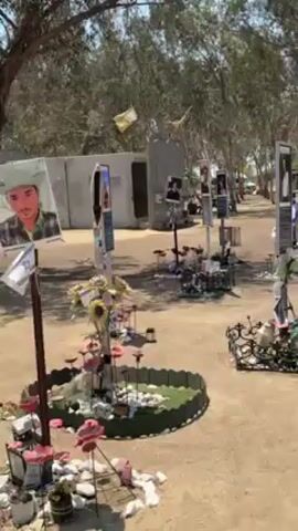The video shows a memorial site with numerous displays set up on sandy ground under a clear blue sky. These displays feature photographs of individuals, accompanied by text on placards. The ground around each display is decorated with various items, including stones, flowers, and small decorative objects. People are seen walking among the displays, some holding photographs or looking at the placards. White buses are visible in the background. The scene appears to be a memorial dedicated to individuals, with visitors paying their respects.
