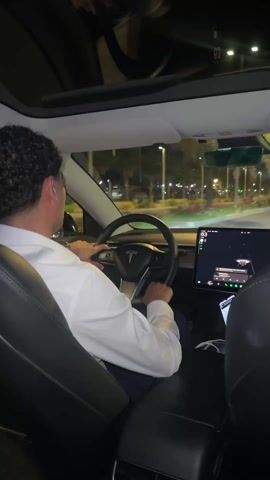The video shows a woman riding in the back of a Tesla at night. The driver is wearing a white shirt and holding the steering wheel. The woman has long dark hair and is wearing a black leather jacket. She is holding a sparkly phone case. She yawns and gestures with her hands, seemingly reacting to the music playing. The car is moving on a brightly lit road.