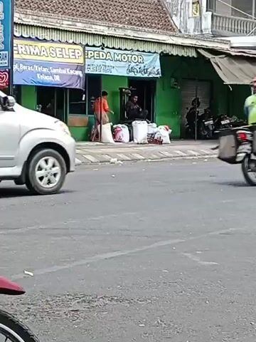A silver minivan drives on a street. On the sidewalk, there are several people and sacks of goods in front of a green building. A motorcycle with two people on it drives past the minivan.