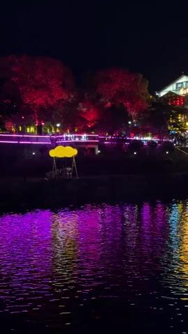 At night, the scene showcases a body of water reflecting colorful lights from buildings and trees along the opposite bank. The water's surface is rippled, creating a distorted reflection of the vibrant colors—primarily pink, purple, and yellow. On the bank, trees are lit in red, purple, and blue.  A yellow cloud-shaped swing set is visible.  People can be seen near the swing set, appearing as small figures against the backdrop of illuminated trees and architecture. The overall atmosphere is peaceful and festive due to the colorful lights.