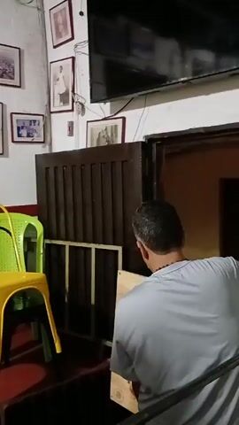 Here is a description of the video:

The video shows a man in a light blue shirt moving a wooden crate in a room with red tile flooring. A yellow chair is visible next to him. The walls are white and adorned with framed pictures. 

Next, another man wearing a bright green shirt and a baseball cap enters the frame and picks up a black metal table frame. He lifts it over his head and carries it through a doorway. A third man, dressed in a vest and jeans, walks past him. The green-shirted man continues carrying the table frame, and the video ends.