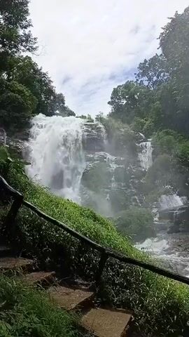 A vertical video shows a powerful, multi-tiered waterfall cascading down a rocky cliff face surrounded by lush green foliage and trees. A wooden railing and a set of stone steps are visible in the foreground, suggesting a pathway for visitors. The sky is partly cloudy. The water is churning and white, creating a mist that rises from the base of the falls. The scene conveys a sense of natural beauty and the raw power of water.