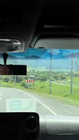 The video is shot from inside a vehicle, looking out the windshield. The road curves to the left, with a guardrail on the right side. Beyond the guardrail is a lush green valley with trees and some buildings. In the distance, mountains are visible under a cloudy sky. A person's head is reflected in the rearview mirror. There are no other people clearly visible in the frame. The interior of the vehicle, including the dashboard and part of the ceiling, is also visible. The time stamp indicates the video is about 4 seconds long.