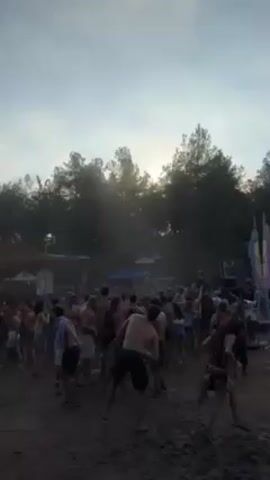 The video shows a large crowd of people dancing at an outdoor event. Many people are shirtless. The ground appears to be dirt. There are trees in the background, and the sky is overcast with the sun visible through the trees. There are tents and speakers visible in the background.