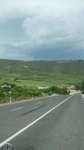 The video captures a first-person perspective from a vehicle driving on a two-lane asphalt road in a rural or semi-rural setting. The road features clear white lane markings and is flanked by green vegetation, including trees and bushes on both sides.

In the background, rolling green hills dominate the landscape, extending towards a distant horizon. Patches of lighter green suggest agricultural fields or open grassy areas. Scattered small buildings or structures can be seen nestled among the hills in the distance. The sky is largely overcast with prominent gray and white clouds, indicating a cloudy but possibly bright day.

The vehicle is moving forward, and the road appears to curve gently in the distance. Small white marker posts are visible along the edge of the road on the right side. In the bottom-left corner of the frame, a Mercedes-Benz logo is partially visible, indicating the make of the vehicle from which the video is being recorded.

The scene conveys a sense of travel through a verdant, hilly landscape under a cloudy sky. The location context provided is Vardashat, Armenia. There is no visible text within the scene itself, apart from the car's logo.