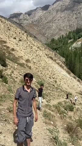 The video begins with a medium shot of a young man standing on a dirt path in a mountainous, arid environment. He is wearing a striped t-shirt, gray sweatpants, and sunglasses, and he is smiling at the camera. Behind him, another young man is walking up the path. The camera then pans to the right, revealing a valley with lush green trees and a village nestled on the hillside. The village consists of many stone and mud-brick houses built close together, characteristic of traditional mountain settlements. The houses are terraced up the slope, with narrow pathways connecting them. Tall, slender trees are interspersed among the buildings, and the backdrop is dominated by a rugged, imposing mountain range. The sunlight is bright, casting shadows and highlighting the textures of the landscape and the village architecture. Several other figures can be seen in the distance, also on the path, suggesting a group is hiking or exploring the area. The overall scene conveys a sense of natural beauty, rural life, and a remote mountain community.