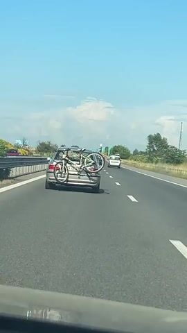 A silver car with a bicycle rack attached to its rear, carrying two bicycles, drives on a multi-lane highway on a clear day. The highway has white dashed lines separating the lanes. There is a guardrail and green vegetation to the left of the highway. Another vehicle, possibly a white van, is visible in the distance in the right lane. The sky is blue with some scattered white clouds.