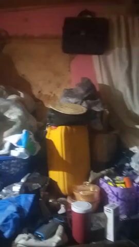 The video depicts a cluttered room with various items piled up. Several bags, baskets, containers, and kitchenware are visible. A yellow jerrycan and a black briefcase add to the disarray. The room has a red-painted wall. The lighting is poor, casting shadows and making it difficult to discern specific details. The camera moves slightly, showing more of the chaotic accumulation.