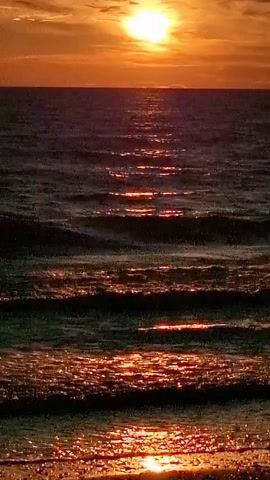 The video shows a sunset over the ocean. The sun is a bright orange ball in the sky, and its rays are reflecting off the water, creating a shimmering path of light. The waves are small and gentle, and the water is a dark blue color. The overall mood of the video is peaceful and serene.