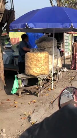 The video shows an outdoor food stall on a sunny day. A vendor, a person of East Asian descent with dark hair and wearing a blue shirt and dark pants, stands behind a table laden with large bags of fried chips. One bag, made of clear plastic, is prominently displayed on the table and filled with golden-brown, circular chips. Behind it, another, larger bag of chips is visible. To the right of the table, a woman wearing a patterned dress and a headscarf is standing. The stall is covered by a large blue umbrella, which provides shade. The ground is dirt and gravel, with scattered dry leaves. A tree trunk is visible on the left side of the frame. Part of a red motorcycle, with a white sticker depicting a dog's face on its side mirror, is in the foreground, suggesting the video is filmed from the perspective of someone on a motorcycle. A sign with Indonesian text is visible on the stall. The overall scene depicts a typical roadside food vendor in what appears to be a warm climate.