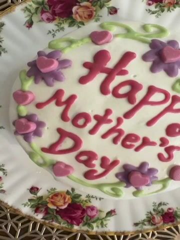 A top-down shot shows a white cake with pink writing that says "Happy Mother's Day" and is decorated with purple flowers, pink hearts, and green vines. The cake sits on a floral-patterned plate, which is placed on a gold-colored patterned surface. The camera slowly zooms out to give a wider view of the cake and its surroundings.