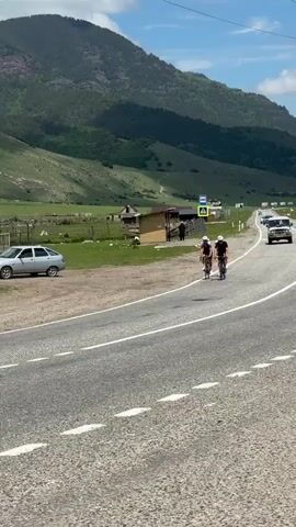 Four cyclists ride in a line on a paved road with white dashed lines. The cyclists are wearing helmets and athletic clothing. One cyclist in the front is waving. The road curves to the right. In the background, there is a green field with some trees and buildings. Beyond the field, there are mountains with a blue sky and some clouds above. A silver car is parked on the left side of the road. A white car is driving in the distance on the right side of the road. A bus stop sign is visible near the road.