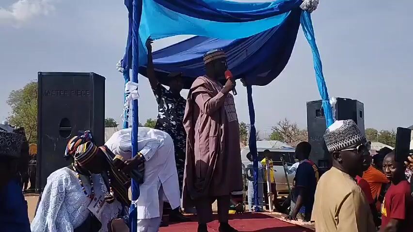 Here's a forensic description of the scene:

**Setting:** An outdoor public event, likely in a town or rural area, given the open space and visible buildings in the background.  The weather appears to be sunny and clear.

**People:**
*   **Central Figure:** A man in a long, brown traditional robe and cap, holding a red microphone. He appears to be speaking to the crowd.
*   **Attendees:** A diverse group of people surrounding the stage. Many men are wearing traditional Muslim caps. One person in a white robe bows low in front of the man speaking. There is a uniformed police officer behind the speaker.

**Objects:**
*   **Stage:** The man is standing on a makeshift stage, indicated by a red carpet on the ground.
*   **Decor:** A blue and white fabric canopy is suspended above the stage.
*   **Speakers:** Two large black speakers labeled "MASTER PIECE" flank the stage.
*   **Microphone:** The man is using a red handheld microphone.

**Actions/Interactions:**
*   The man in the robe is addressing the crowd with a microphone.
*   The person in the white robe is bowing.
*   Other individuals are listening and observing.

**Environment:** The background shows a relatively flat landscape with scattered trees and buildings. The sky is a clear blue.