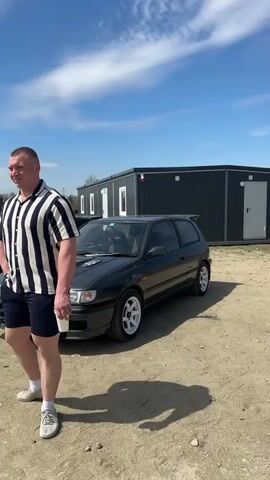 The video shows a man walking by a black Nissan Pulsar car with white rims.  The video then pans right to show a trailer with its back doors open, revealing large speakers on the doors and inside the trailer. Three men are near the trailer: one in army fatigues, one in jeans and a black shirt, and one inside the trailer who appears to be DJing. The environment appears to be a dirt lot with a clear, blue sky above. There are gray storage containers in the background.