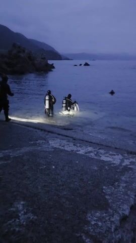 The video shows four divers with scuba gear at the edge of a body of water at dusk or dawn. One diver stands on a concrete ledge, while the other three are in the water, with two close together. They are using lights, possibly to illuminate the water.  Mountains are visible in the background under an overcast sky.