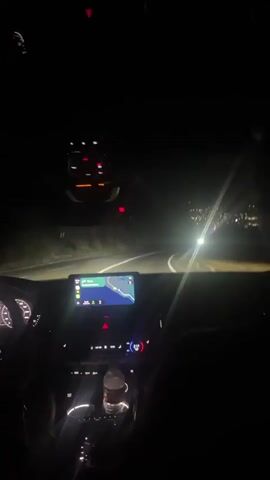 The video is shot from the perspective of a passenger in a car at night. The dashboard is visible in the foreground, with a navigation screen displaying a map and a car icon. The driver's hand is on the steering wheel on the left side of the frame. The road ahead is dark, with oncoming headlights visible. In the distance, a city or town's lights can be seen on a hillside. The car appears to be moving along a winding road.