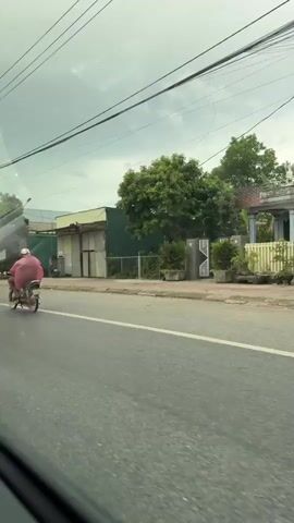 A person wearing a pink poncho and a white helmet rides a motorcycle on a paved road. The person is large and appears to be wearing loose-fitting clothing under the poncho. The motorcycle is brown and appears to be an older model. The road is lined with buildings and trees. The sky is overcast. The video appears to have been filmed from another vehicle.