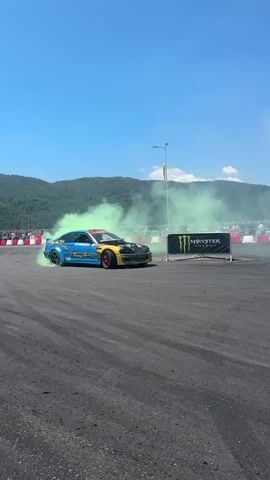 A blue and yellow drift car is shown performing a drift on a race track. The car is emitting a large cloud of green smoke from its tires. In the background, there are spectators behind a barrier, mountains, and a bright blue sky. A banner for Monster Energy is visible behind the car. The car's wheels are turned sharply, indicating it is in the middle of a drift. The driver is visible inside the car. The track appears to be made of asphalt.
