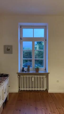 The video pans across a room with cream-colored walls and hardwood floors. The first shot shows a large white radiator below a window with four panes. The window looks out onto greenery, and a small framed picture hangs on the wall to the left. Next, the camera pans to the left, revealing a dresser with a marble top, a television, and two decorative arched shelves, each containing a small red lamp. A tall, decorative object hangs on the wall in the next shot. The camera then moves to the right, showing a gray armchair with luggage on it, a bookcase filled with books, and a large window with gray curtains. The final shot shows two decorative palm tree lamps standing on a red rug, with the bookcase and armchair visible in the background.