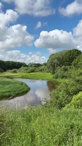 The video opens with a wide shot of a serene natural landscape. A calm body of water, possibly a river or lake, snakes through a lush green meadow. The water reflects the bright blue sky dotted with fluffy white clouds. Dense foliage, including trees and bushes, forms a natural backdrop on either side of the meadow and water. The foreground is dominated by tall, vibrant green grass, suggesting a sunny day. The camera slowly pans to the right, following the curve of the water. As the scene progresses, the viewpoint shifts slightly, and two tall, dark, rusty metal structures are visible, seemingly part of some old infrastructure. Next to them, a rough, earthy path is seen, and some weathered concrete structures are present. The vegetation remains dense and green, with a mix of grasses and trees. The sky continues to display a clear blue expanse with scattered clouds. The video ends with a closer view of the path and the concrete structures, with trees and vegetation surrounding them. There are no people visible in the video. The overall impression is one of a peaceful, overgrown, and somewhat abandoned rural or natural environment.
