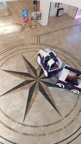 This is a high-angle, forward-moving video shot inside a large, well-lit indoor space with marble floors and a large compass rose design. In the center of the frame is a toy train with multiple carriages, each painted white and dark blue with red accents. The train is moving in a counter-clockwise direction along the compass rose. A person in dark clothing is walking away from the train in the upper right quadrant of the frame. In the background, there are several information display stands, a fire extinguisher, and a white pillar. The train's lights are visible and illuminated.