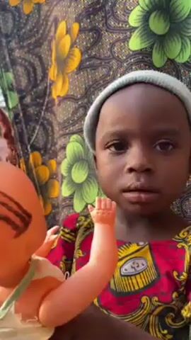 A young, dark-skinned child with a pale-green beanie is holding a baby doll. The child is wearing a red and yellow patterned shirt and is standing in front of a patterned yellow, green, and black backdrop. The doll is orange and has brown yarn braids, with a white and green top. The child looks at the doll and then looks to the side.