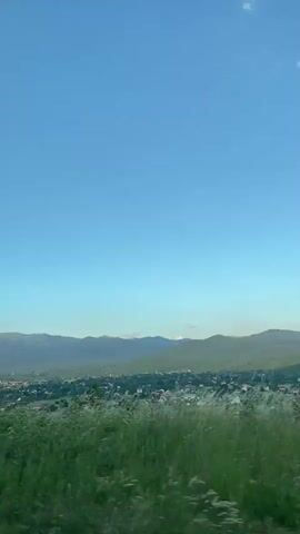 A vertical video captures a panoramic view of a rural village nestled amidst rolling green hills under a vast, clear blue sky with a few wispy clouds. In the foreground, the edge of a paved road is visible, bordered by tall, dry grass and some green foliage. As the camera pans slightly to the right, the village comes into clearer view, with houses and buildings scattered across the landscape. The distant mountains form a dramatic backdrop, their slopes covered in lush greenery. The lighting suggests it is either early morning or late afternoon, with a soft, natural light illuminating the scene. There are no people or animals visible in the video.