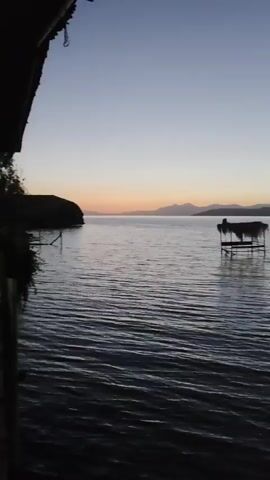 The video captures a wide shot of a calm body of water, likely a lake or sea, at either dawn or dusk. The sky is a gradient of pale blue fading to a soft orange near the horizon. In the distance, a silhouetted mountain range is visible. A small, dark object, possibly a boat or raft, floats on the water in the mid-ground. The water itself is dark and exhibits gentle ripples. The foreground is darker, suggesting the camera is positioned at a low angle, possibly from a dock or a boat. There are no visible people or discernible actions taking place. The overall atmosphere is serene and tranquil.