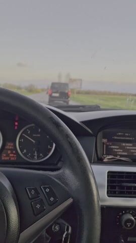 The video shows a driver's perspective from inside a BMW. The steering wheel is in focus, with gauges and a central console visible. The dashboard displays information, including time (7:17), temperature, and odometer readings. The radio is tuned to 107.8 MHz. Outside the car, the road stretches ahead with sparse greenery on the sides. Other vehicles are visible in the distance. The sky is overcast, suggesting either early morning or late afternoon. The car is in motion.