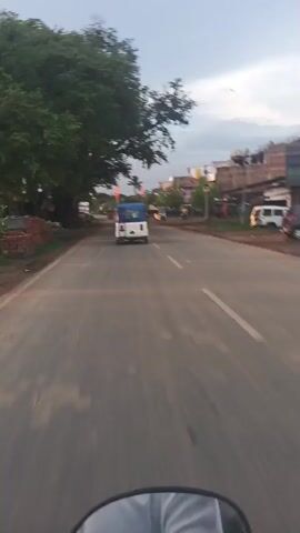 The video shows a view from a moving vehicle, possibly a motorcycle, traveling down a two-lane road. In the distance, an auto-rickshaw with a blue canopy is visible in the center of the frame. To the right of the road, there are buildings with shops and some parked vehicles. A large tree with dense foliage is on the left side of the road, and its branches extend over the roadway. The sky is cloudy, suggesting either early morning or late afternoon. There are also some other vehicles and people on the road and sidewalks, but they are not the primary focus. The road appears to be paved and has lane markings. The overall scene depicts a typical roadside environment in a town or city.