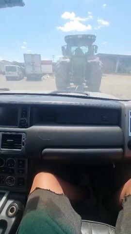 The video, filmed from the interior of a vehicle, shows the dashboard and the legs of a person wearing denim shorts and sneakers. Through the windshield, a large green tractor is visible from the rear, its tires large and its engine running. To the left of the tractor, a white trailer is parked. The environment appears to be an outdoor area, possibly a farm or industrial yard, with a bright, sunny sky overhead. The tractor is stationary at the beginning of the clip but appears to start moving slightly as the video progresses.