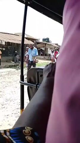 The video captures a lively daytime street scene in Obunta-Isiala, Nigeria, from the perspective of someone inside a vehicle, likely a tricycle (Keke Napep). The camera is positioned looking out from the side, with a dark-skinned arm, possibly the driver's, prominently in the foreground, resting on the vehicle's frame. A blurry purple fabric is visible to the right.

The setting appears to be an unpaved or dirt road, indicative of a local street or market area, lined with simple buildings featuring corrugated iron roofs. The bright sunlight suggests clear weather.

Numerous pedestrians are seen walking past the vehicle. Individuals include a man in a black t-shirt and blue shorts, another man wearing a red and white football jersey, and a woman in a black t-shirt with the letters "OK" visible on it. At one point, a man in a white t-shirt briefly leans over towards the vehicle's side before moving on. In the background, a large billboard with the text "UCHENNA CHERIBE" is partially visible. The scene depicts everyday activity in a bustling environment. Music can be heard playing throughout the video.