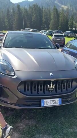 A close-up, front-facing shot of a gray Maserati SUV parked outdoors on a grassy area. The vehicle has a distinctive trident logo on the grille and an Italian license plate. In the background, a parking lot filled with various vehicles is visible, backed by a dense forest of pine trees and a mountain slope. The sky is clear and blue with a single cloud. The camera then pans slightly to the left, showing the side of the car and the headlights. It then moves to a wider shot, revealing more of the parking lot and the surrounding mountainous, forested landscape. The video is taken during the daytime.