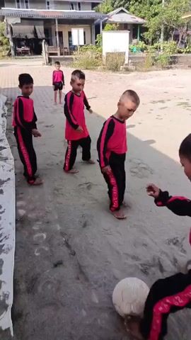 A group of young boys, likely primary school-aged, are actively playing a game outdoors on a dusty, sandy ground. They are all uniformly dressed in matching red and black long-sleeved athletic tops and black track pants with red stripes. Most of the boys are barefoot.

The central activity involves them kicking a small, light-colored, spherical object (resembling a stone or a worn ball) amongst themselves and against a white tarp-like structure positioned to their left. The game appears to be an informal variation of soccer or kickball, with the boys using their feet to keep the object in motion. Another boy can be seen standing further in the background, observing the play.

A notable detail is the text "UPTD SD NEGERI 14 JULI KAB. BIREUEN" clearly visible on the back of one boy's uniform, identifying their school and location as Kabupaten Bireuen, Indonesia.

The scene is set in what appears to be a rural or semi-urban environment, with houses and green foliage visible in the background under clear, bright daylight, suggesting it is either morning or afternoon. The boys appear focused and engaged in their play.