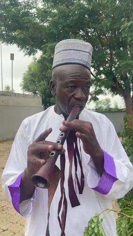 A man wearing a white robe and a patterned cap plays a dark wooden wind instrument outdoors. The instrument has a flared bell and several dark ribbons hanging from it. The man is holding the instrument with both hands and blowing into it. In the background, there are trees, a wall, and a light pole.