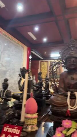 The video pans across a dimly lit interior, likely a temple or shrine, showcasing various statues of Buddhist figures. The statues appear to be made of dark wood or metal, with intricate details and ornate decorations. In the foreground, a seated Buddha statue is prominently displayed, adorned with a white beaded necklace. To the right and behind it, a much taller, standing statue is visible. The ceiling is dark wood with recessed lighting, and the walls are decorated with murals and fabric drapery. The atmosphere is serene and reverent, with a focus on the religious artifacts.