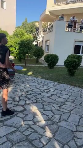 Two young males are playing paddle ball in a courtyard. One male is in the foreground facing away from the camera, wearing black shorts and no shirt. He is holding a paddle in his right hand, poised to strike a ball. His left hand is raised, possibly having just thrown or served the ball. The other male is in the midground, facing the foreground male, wearing grey shorts and a black and grey t-shirt. He is crouched low, holding a paddle, and appears to be mid-swing or preparing to hit a ball. In the background, on a balcony of a multi-story building, three adult males are standing and observing the game. The environment is sunny with a clear blue sky. The ground is paved with irregular stones, and there is a grassy area with bushes. A parked car is partially visible to the left of the foreground male.