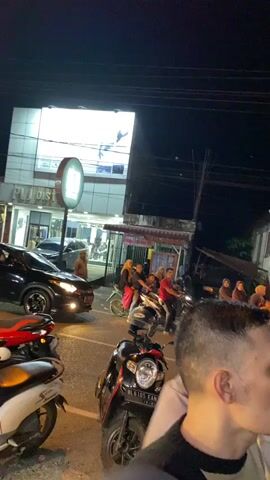 The video shows a busy street scene at night. There are many people gathered, some on motorcycles and some on foot. Buildings with illuminated signs line the street. Several motorcycles are parked or in motion. A white car is visible in the foreground. The overall impression is of a lively gathering or event in an urban setting at night.