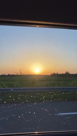 The video begins with a view of a sunset over a field, the sun a bright orb on the horizon. The camera then pans to a shot of the sun peeking through the leaves of a tree, casting dappled light. Next, a reflection of the sunset is seen on the surface of dark water, creating streaks of light. Finally, the video shows a blurred image of the sunset reflected in what appears to be water or a wet surface, with abstract patterns of light and color.