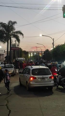 The image captures a bustling street scene at dusk in Lanús Este, Argentina. A large crowd of people, many dressed in red, fills the street and sidewalks, suggesting a gathering or procession. Vehicles are intermingled with the crowd, including a silver Volkswagen hatchback (license plate UHH 150) with its brake lights illuminated in the foreground, a white van, and several motorcycles.

The sky is transitioning from daylight to evening, displaying soft hues of grey, pink, and orange near the horizon, indicating sunset or twilight. Streetlights are on, casting pools of light, and a green traffic light is visible at the top left. Overhead, a network of power lines or cables crisscrosses the sky. Palm trees and other trees line the street, with urban buildings visible in the background.

A large "BRIDGESTONE" sign is prominently displayed on a structure on the left side of the street. A person wearing a black jacket and a yellow shirt is partially visible in the bottom right corner of the frame. The scene conveys a sense of high activity and a considerable number of people participating in or observing an event.