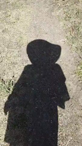 The video shows a shadow of a person wearing a wide-brimmed hat walking on a dirt path. The shadow is cast on the ground, which is partially covered with dry grass and some small rocks. The person's feet are visible at the bottom of the frame at the 00:02 mark, with red painted toenails. The shadow moves from the bottom of the frame upwards and slightly to the right. The lighting suggests it is daytime.