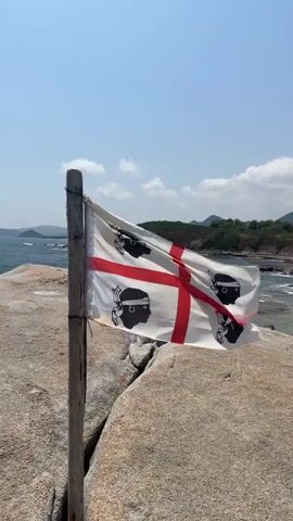 A close-up shot features a flag waving gently in the breeze, attached to a wooden pole. The flag is white with a red cross and four black figures with red bandanas. The pole is positioned on a rocky surface, with the sea and a distant coastline visible in the background. The sky is clear and blue with scattered clouds.
