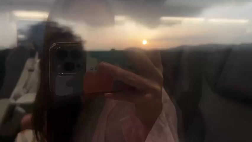 A close-up shot captures a person's hands holding a smartphone in front of a window. The smartphone, with a colorful patterned case and multiple cameras, is positioned to take a selfie. The window reflects the person's face and shoulders, partially obscuring the view outside. Through the window, the sky appears overcast with a bright sun setting or rising, casting a warm glow. The landscape outside is blurred, suggesting motion, possibly from a train or vehicle. The interior of the vehicle, with gray seats, is visible in the background. The overall impression is a travel selfie taken during a scenic moment.