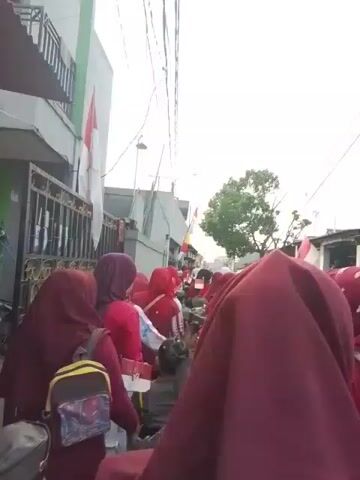 The video shows a group of people walking down a narrow street. The people are mostly women wearing hijabs, and some are carrying flags. The flags appear to be red and white, which are the colors of the Indonesian flag. The buildings on either side of the street are modest, and there are power lines overhead. The sky is overcast. The video is shaky, suggesting it was filmed while the person was walking.